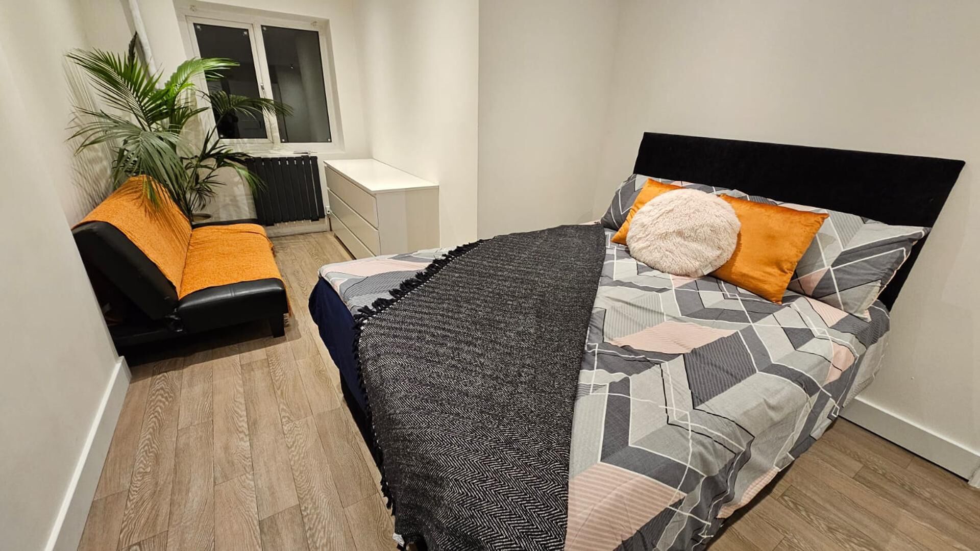 Ein Schlafzimmer mit Bett, Sessel, kleinem Tisch, Topfpflanze und Heizkörper.