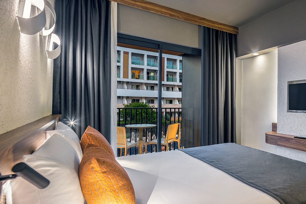 A hotel room with a bed, a TV, a mirror, and a balcony with outdoor seating.
