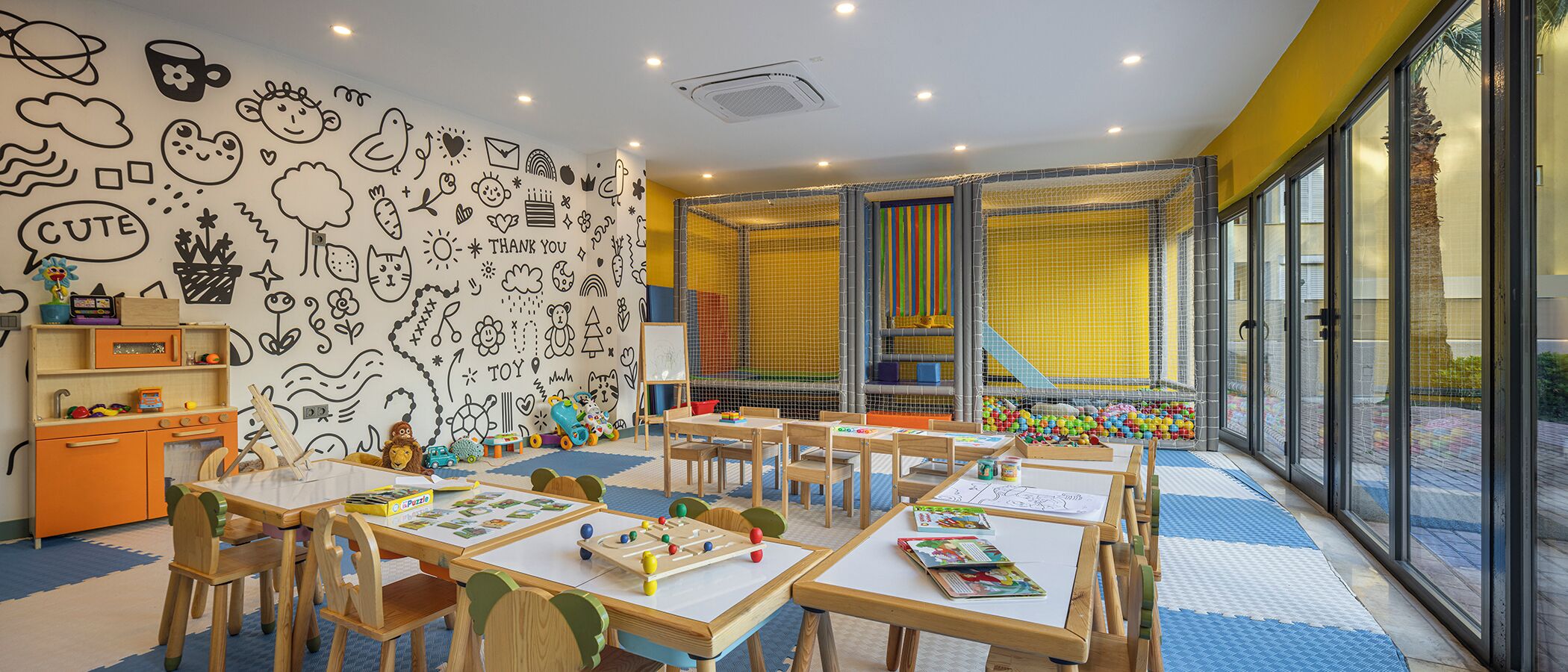 A children's playroom with a wall decorated with doodles, tables with educational materials, and a play area with a net.