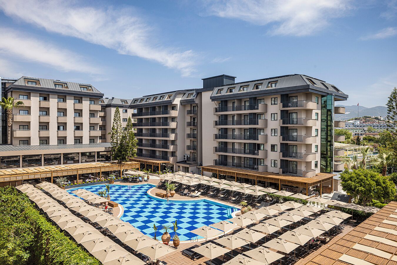 Un hotel con varios pisos, una piscina central con diseño de ajedrez y zonas de estar exteriores con sombrillas.