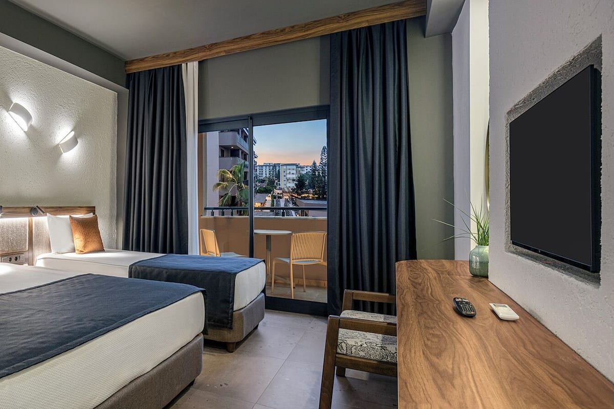 A hotel room with two beds, a desk, a chair, and a balcony with a view of the city.