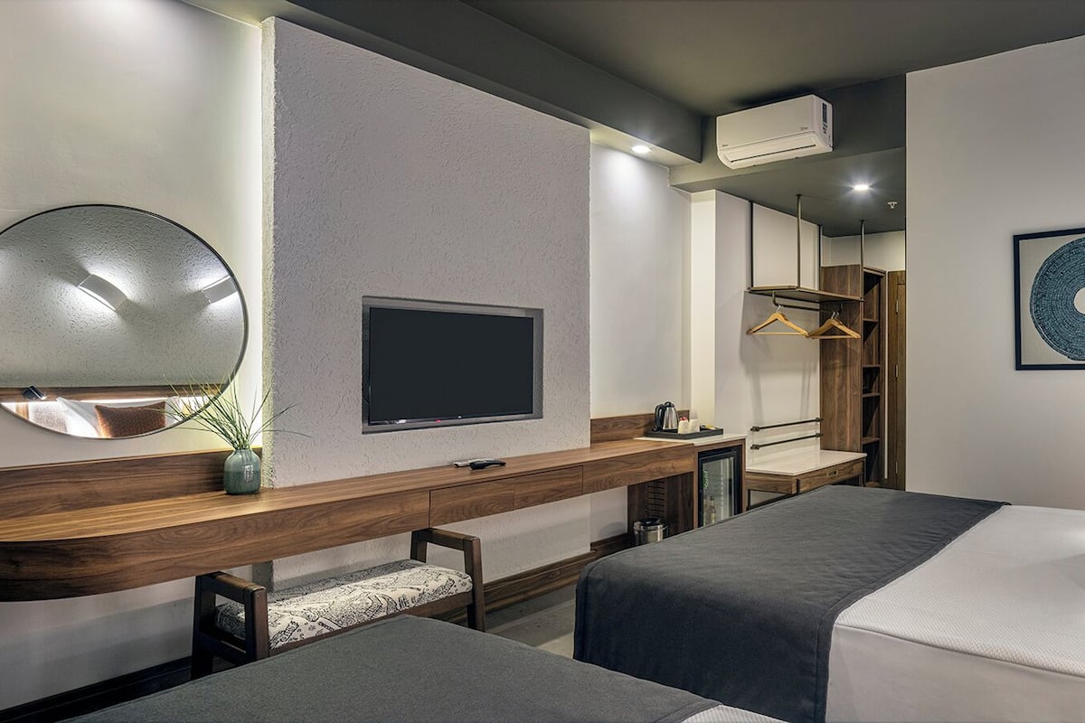A hotel room with a bed, bedside tables, a TV, a mirror, a wardrobe, and a wall-mounted air conditioner.
