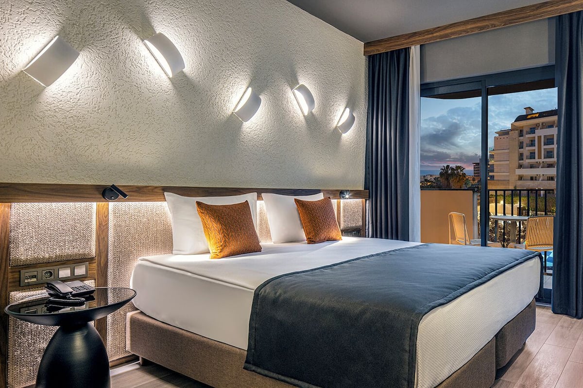 A modern hotel room with a large bed, a small table with a phone and remote controls, a balcony with a view of buildings, and a large mirror on the wall.