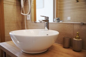 Ein weißes Keramikbecken mit einem modernen Wasserhahn, einer Holzarbeitsplatte, einem kleinen zylindrischen Behälter und einem goldfarbenen Seifenspender.