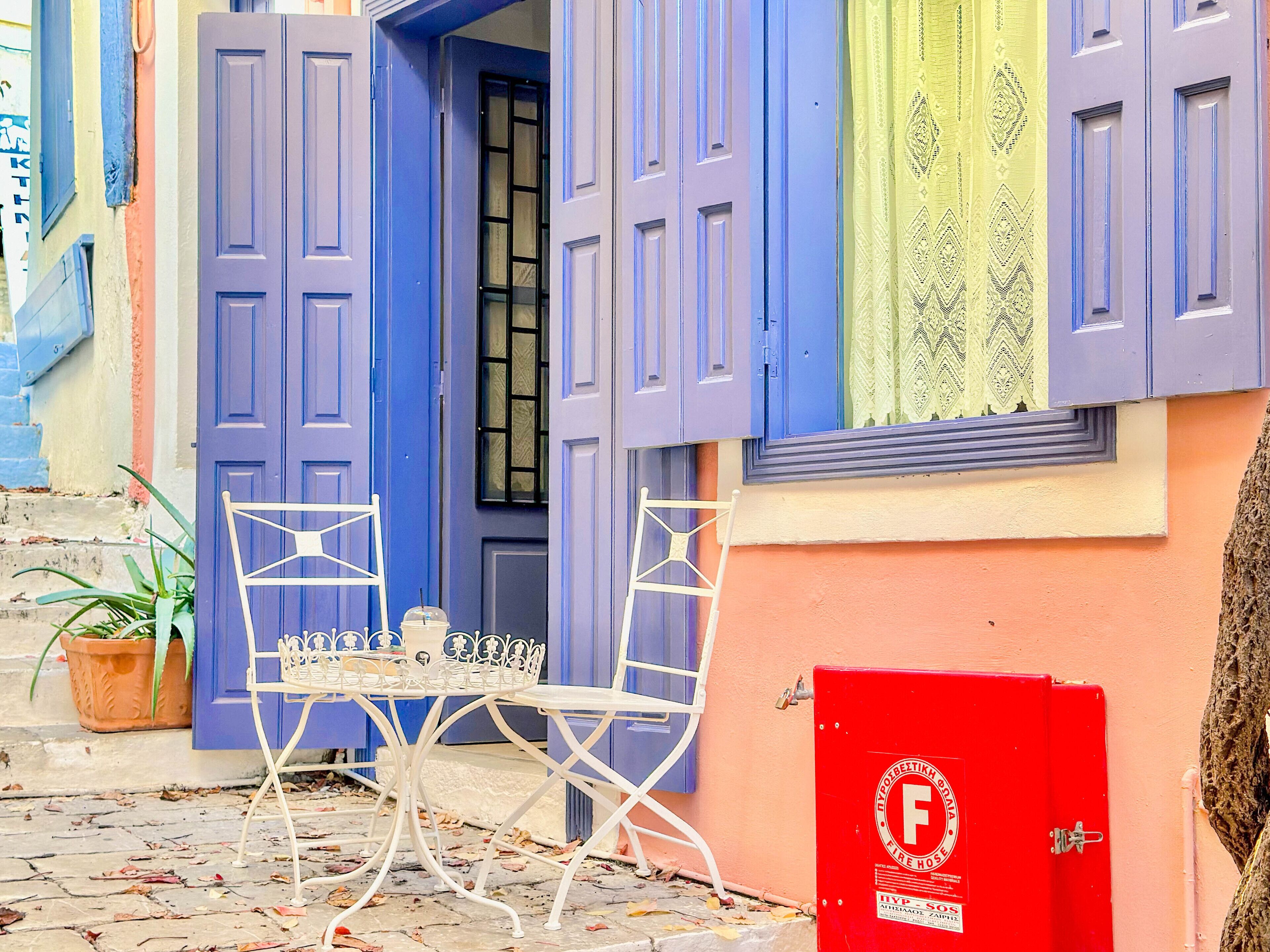 Una escena callejera con una puerta azul, una mesa blanca con sillas y un cuadro rojo para mangueras contra incendios.