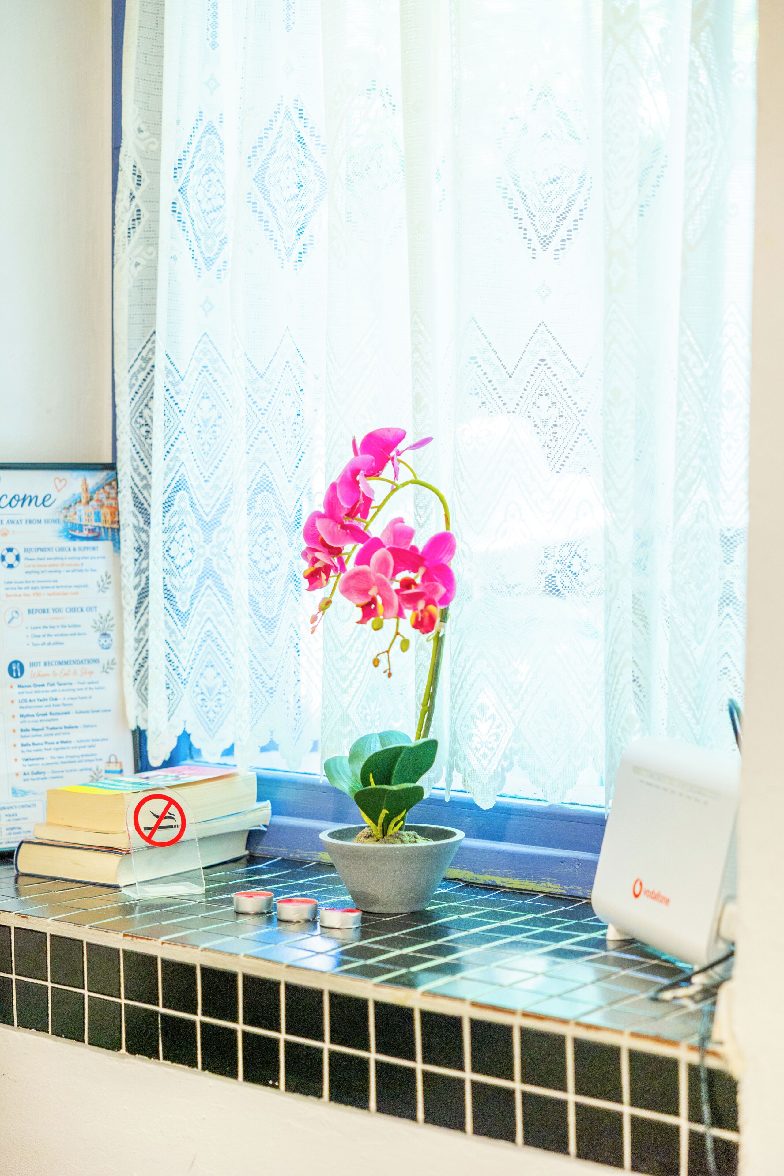 Ein Fenster mit einer Topf-Orchidee, einem Rauchverbot-Schild und einem Vodafone-Gerät auf einer gefliesten Ablagefläche.