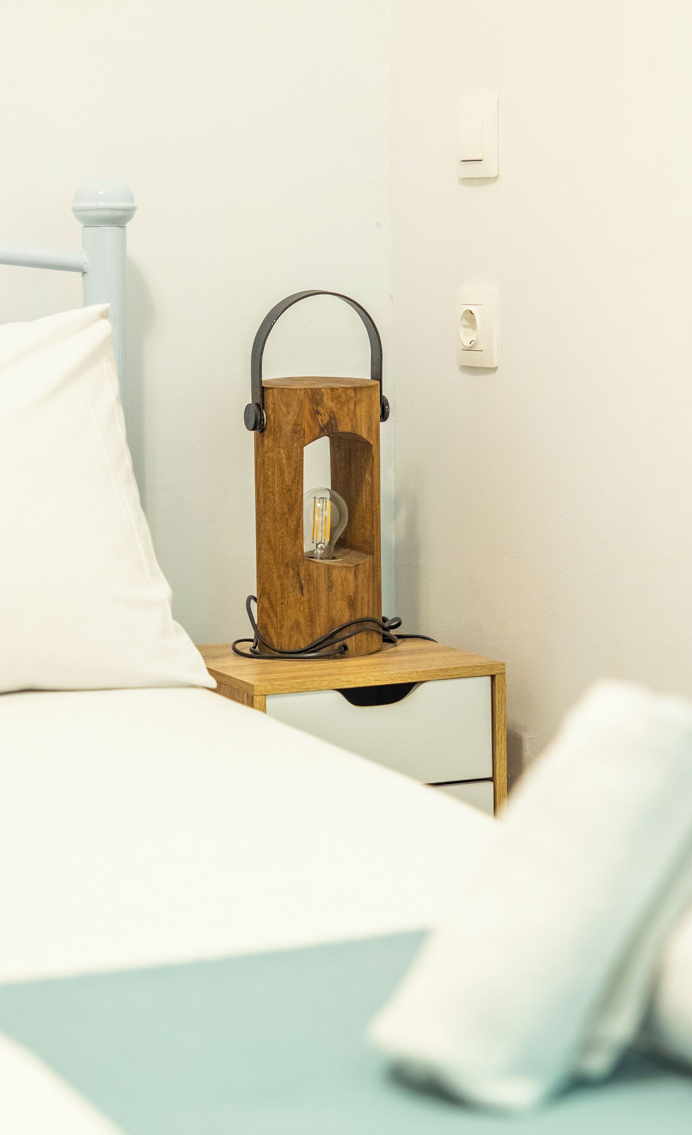 Un lit avec une literie blanche, une table de chevet en bois avec une lampe et une prise électrique au mur.