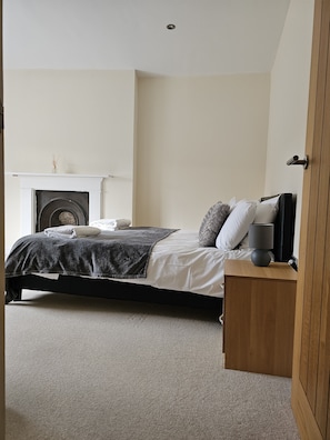 A bedroom with a bed, a nightstand, a lamp, a fireplace, and a wooden door.