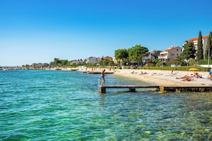 A coastal beach scene with clear water, a pier, and buildings in the background. - Floral Apartments Yazzy 1 (Novalja)
