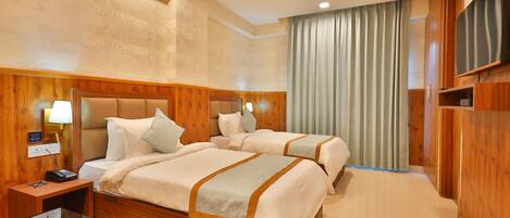 A hotel room with two beds, wooden headboards, a flat-screen TV, a bedside table with a lamp, and a large window with curtains.