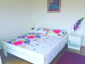 A bed with a floral bedspread, a white nightstand with a lamp, and a purple rug. - Floral Apartments Yazzy 2 (Novalja)