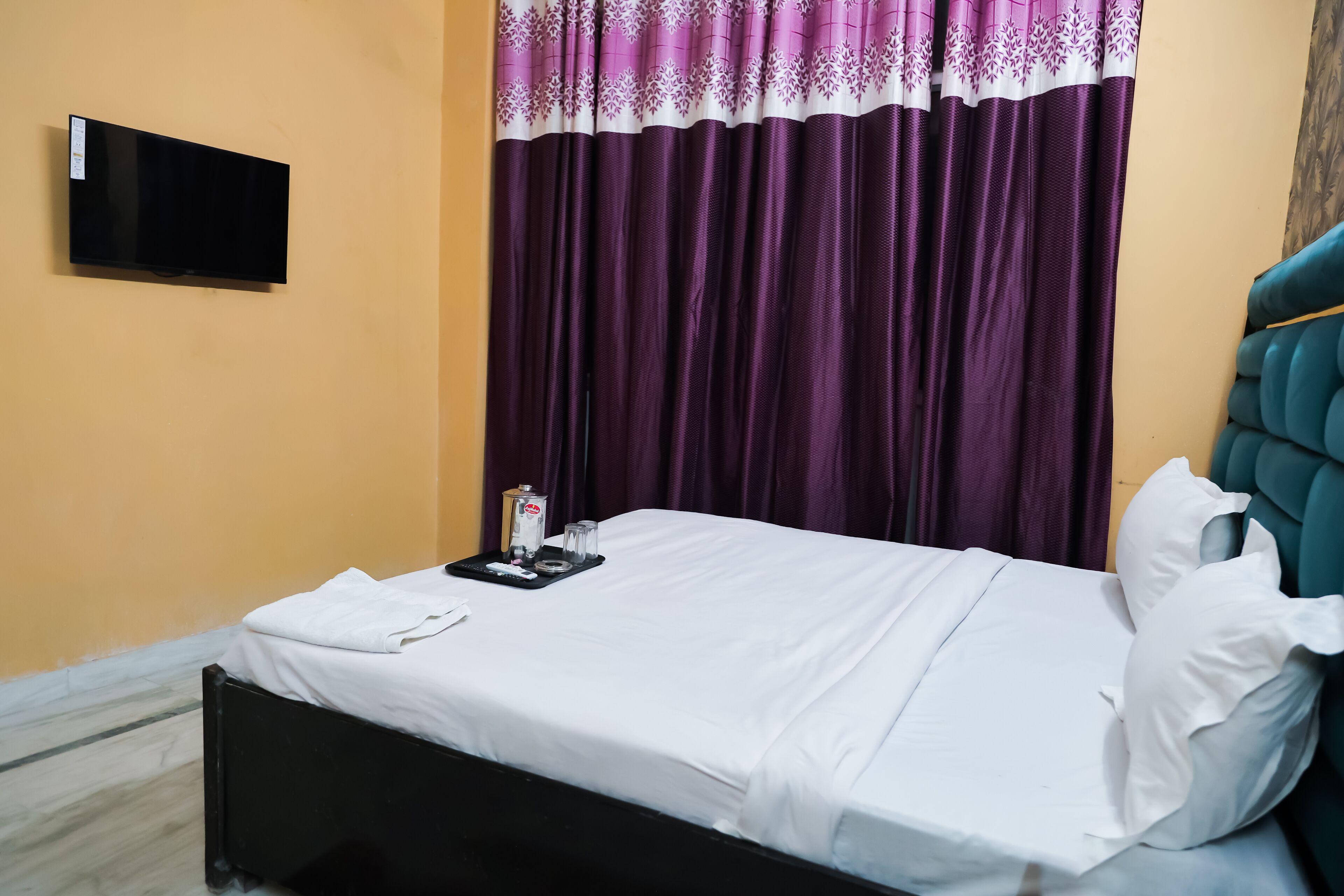 A hotel room with a bed, a flat-screen TV mounted on the wall, and purple curtains.