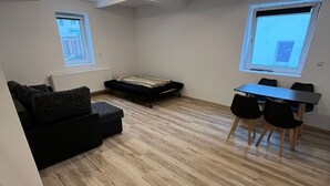 Een knus woongedeelte met een bank, een klein bed, een eettafel met stoelen en een plafond met inbouwspots.