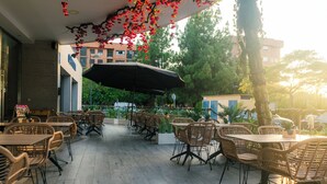 Un espace de café en plein air avec des tables et des chaises, un grand parasol et une vue sur les bâtiments en arrière-plan.
