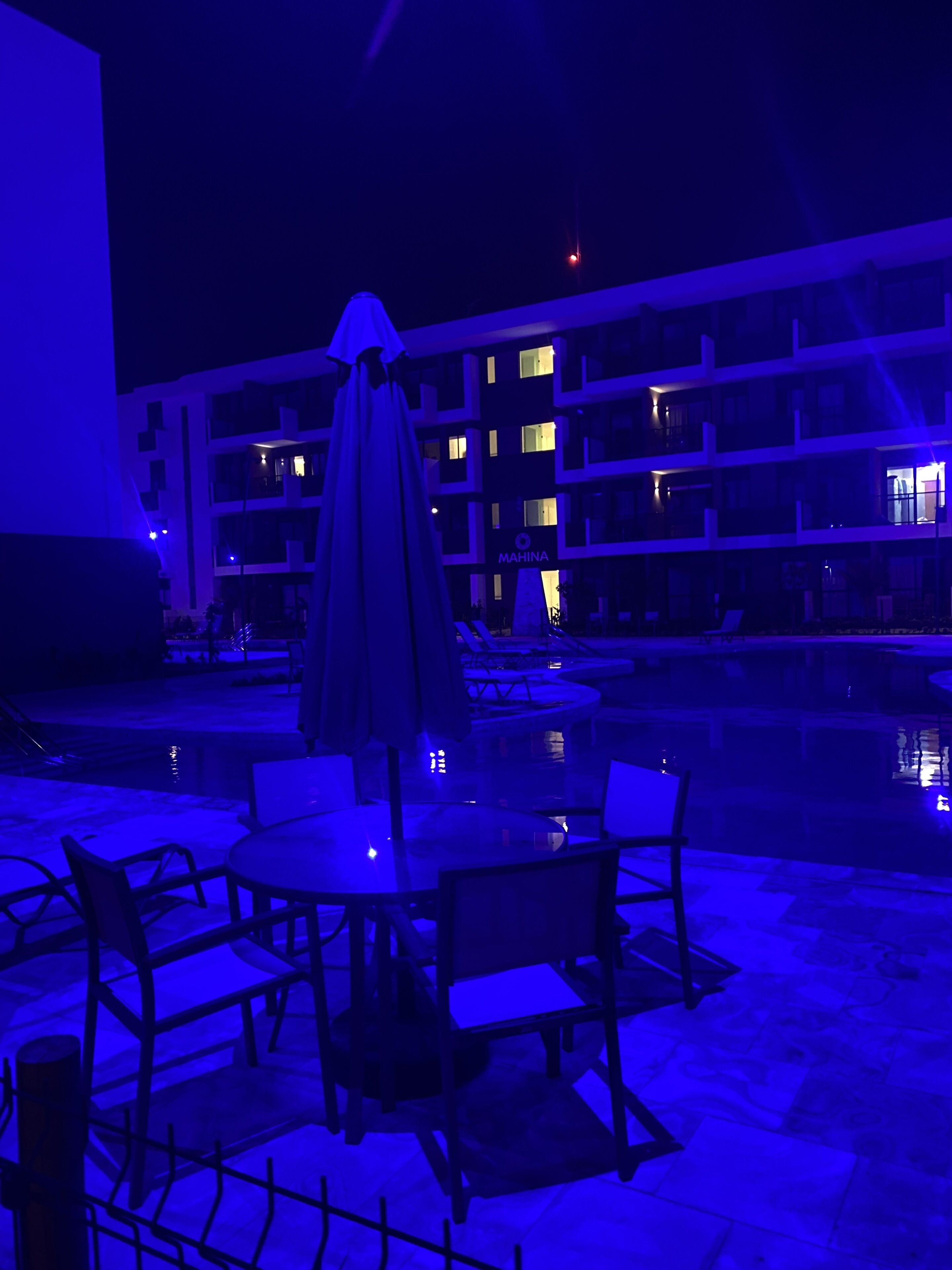 An outdoor pool area with tables and chairs, a covered umbrella, and a building in the background.