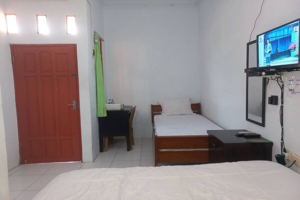 A bedroom with a bed, a desk with a chair, a TV mounted on the wall, and a door.