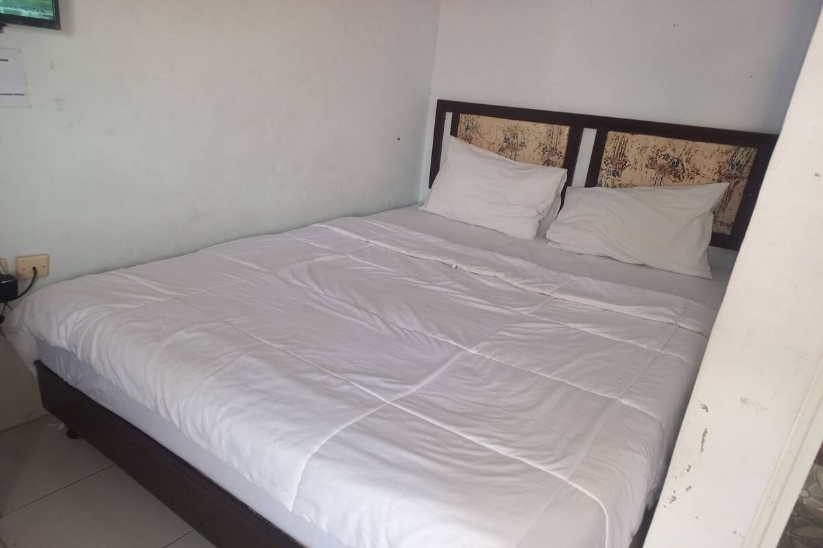 A single bed with a wooden headboard and white bedding in a room with white walls and a tiled floor.