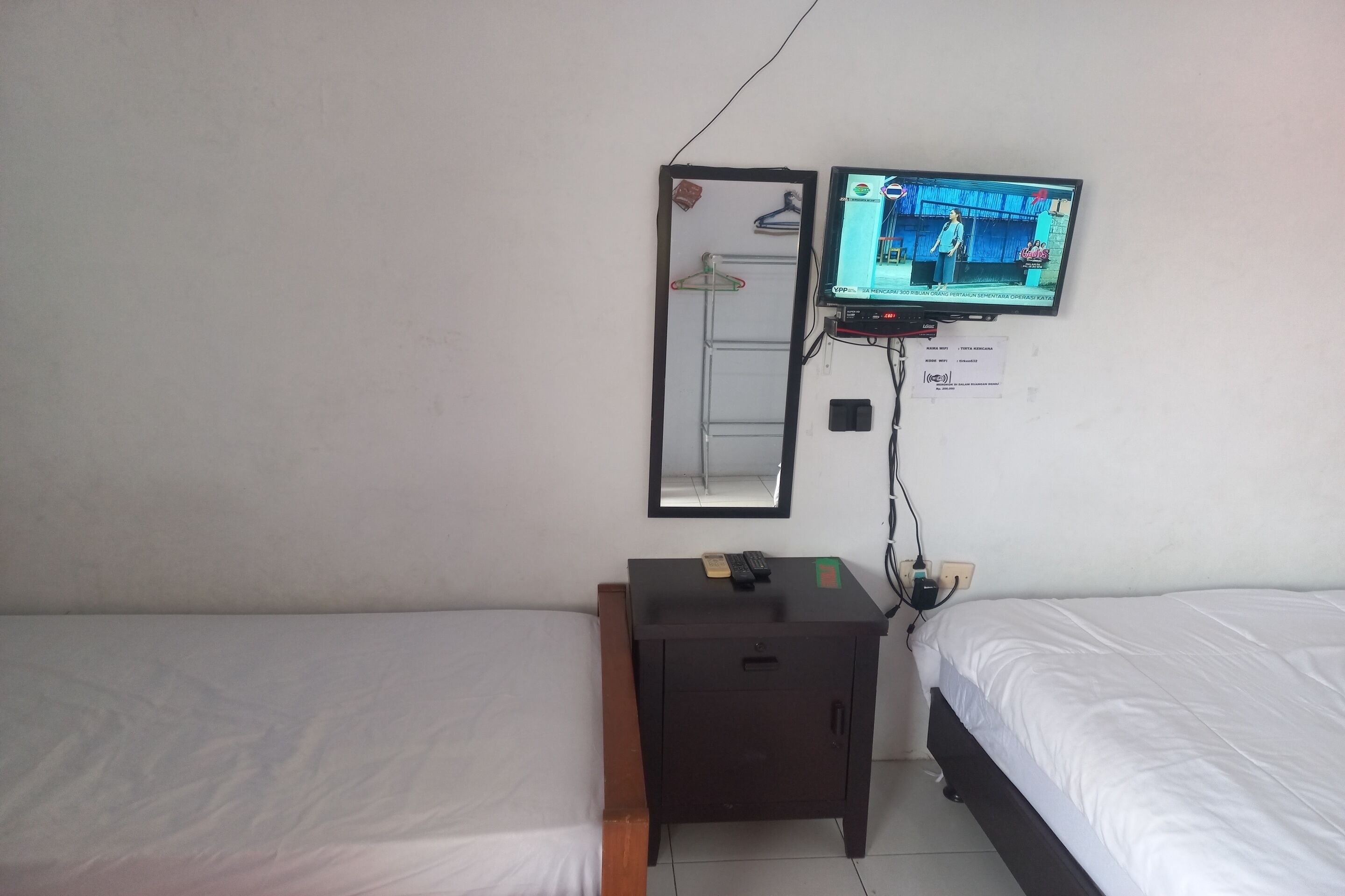 Quarto pequeno e simples com cama, mesa de cabeceira, TV fixada na parede e espelho.