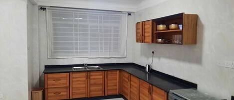 Una cocina con armarios de madera, encimera negra, estufa y ventana con celosías.