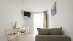 A minimalist living room with a sofa, a dining table with chairs, a flat-screen TV mounted on the wall, and a window with curtains. - Apartment "luka" (Vodice)
