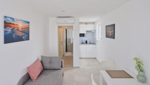 A modern living room with a grey sofa, a small table, and a vase with flowers. A kitchen is visible in the background with white cabinets and a microwave. - Apartment "luka" (Vodice)
