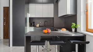 Eine moderne Küche mit einer Marmorarbeitsplatte, einem Spülbecken und einer Obstschale. Auf der Arbeitsplatte stehen zwei schwarze Barhocker und ein Blumentopf.