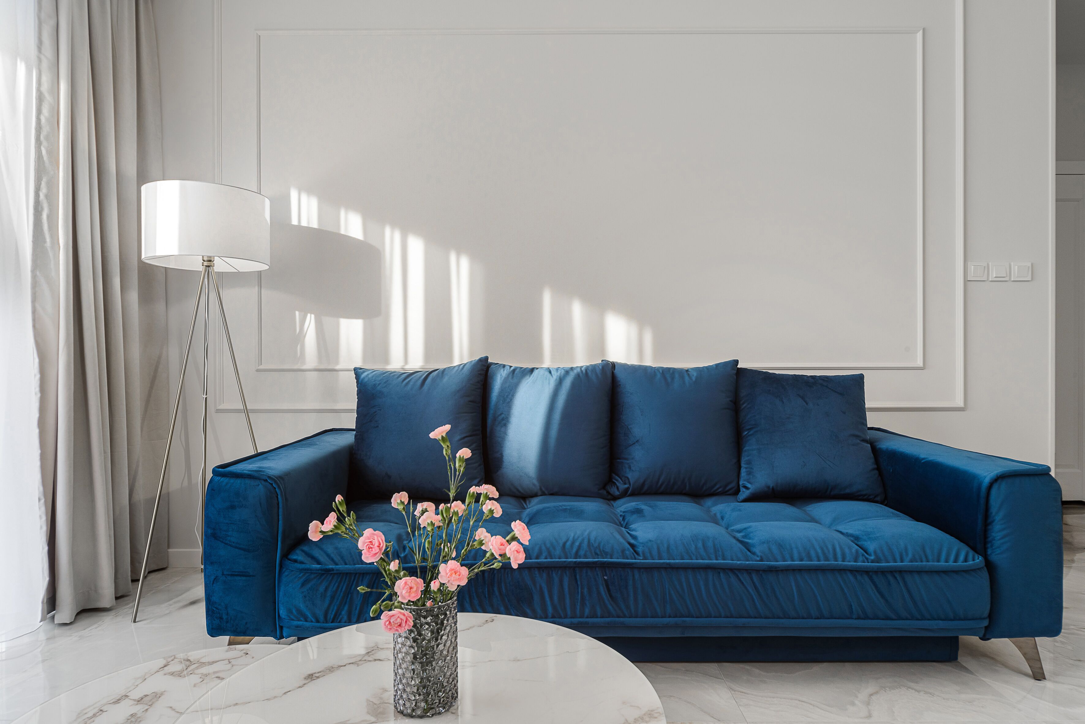 A modern living room with a blue sofa, a floor lamp, and a vase of pink flowers on a marble table.