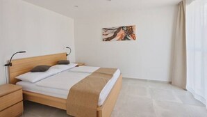 A bedroom with a wooden bed, white bedding, a nightstand, a wall-mounted lamp, and a painting.