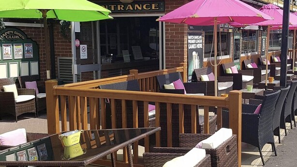 An outdoor seating area with wicker furniture and umbrellas, a brick building with a sign reading "INTRANCE", and a menu board advertising Premier League games. - Beautiful 3-bed Static Caravan in Skegness (Skegness)
