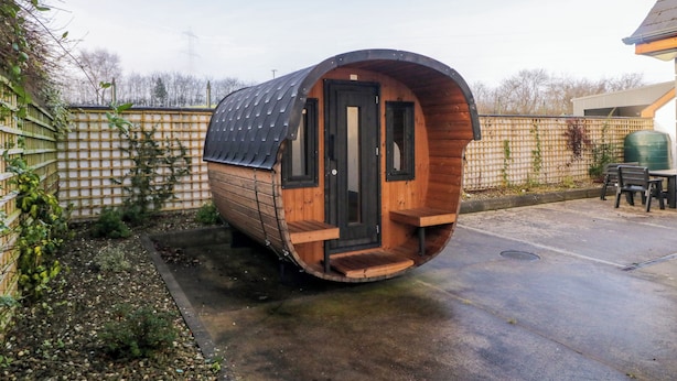 Une structure en bois aux lignes courbes, dotée d’une porte et de fenêtres, située dans un jardin agrémenté d’une clôture et de mobilier d’extérieur.