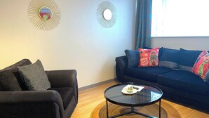 A living room with a dark brown sofa, a round glass coffee table, and a blue sofa with colorful cushions. Two wall-mounted sunburst mirrors are visible. - Impeccable Spacious 6-bed House in Romford (Romford)