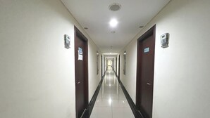 Un long couloir étroit aux murs blancs et au sol recouvert de carreaux noirs. Des portes se trouvent de part et d’autre, certaines étant surmontées d’enseignes.