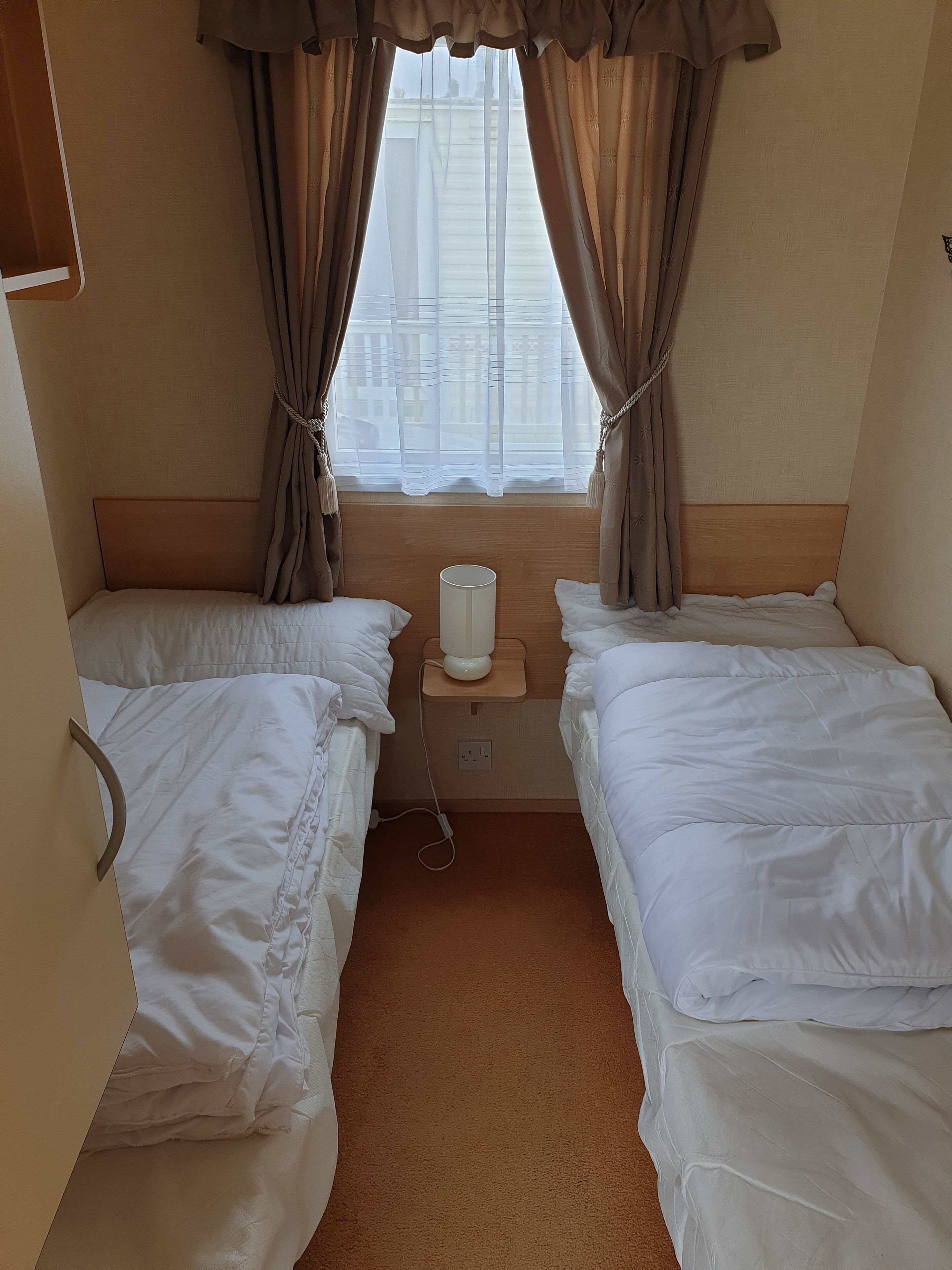 A hotel room with two single beds, a bedside table with a lamp, and a window with curtains.