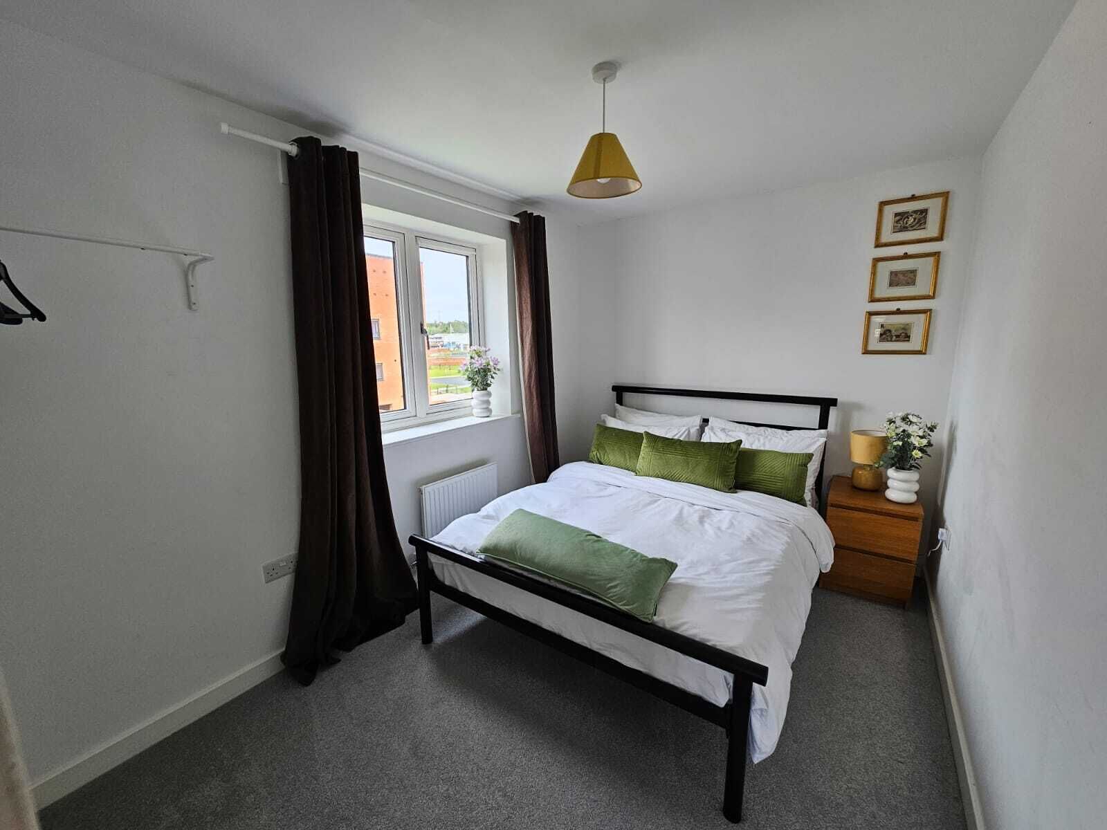 A bedroom with a bed, a window with curtains, a radiator, a bedside table with a lamp, and framed pictures on the wall.