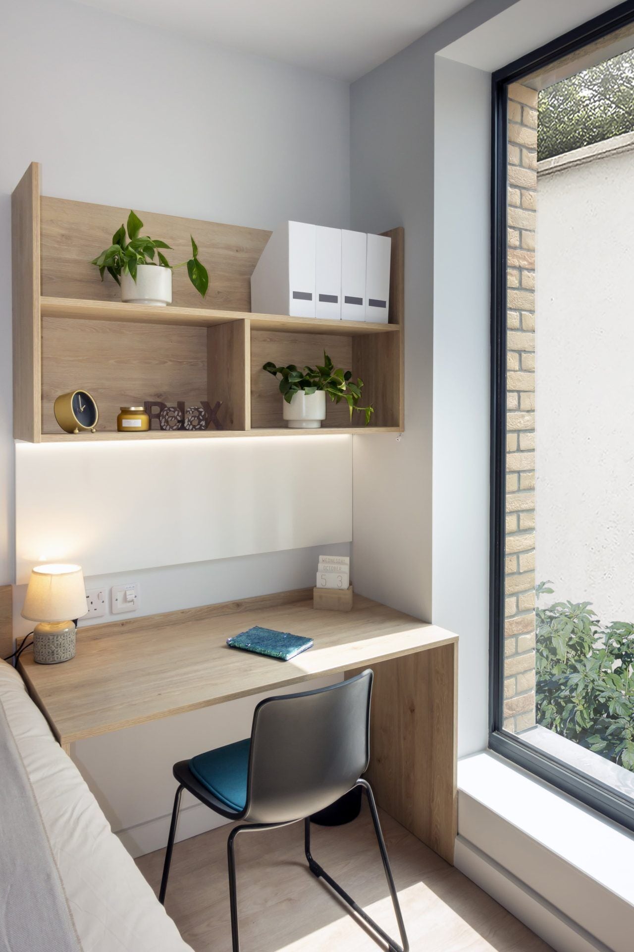 A modern study area with a wooden desk, a chair, a lamp, and shelves with plants and storage boxes.