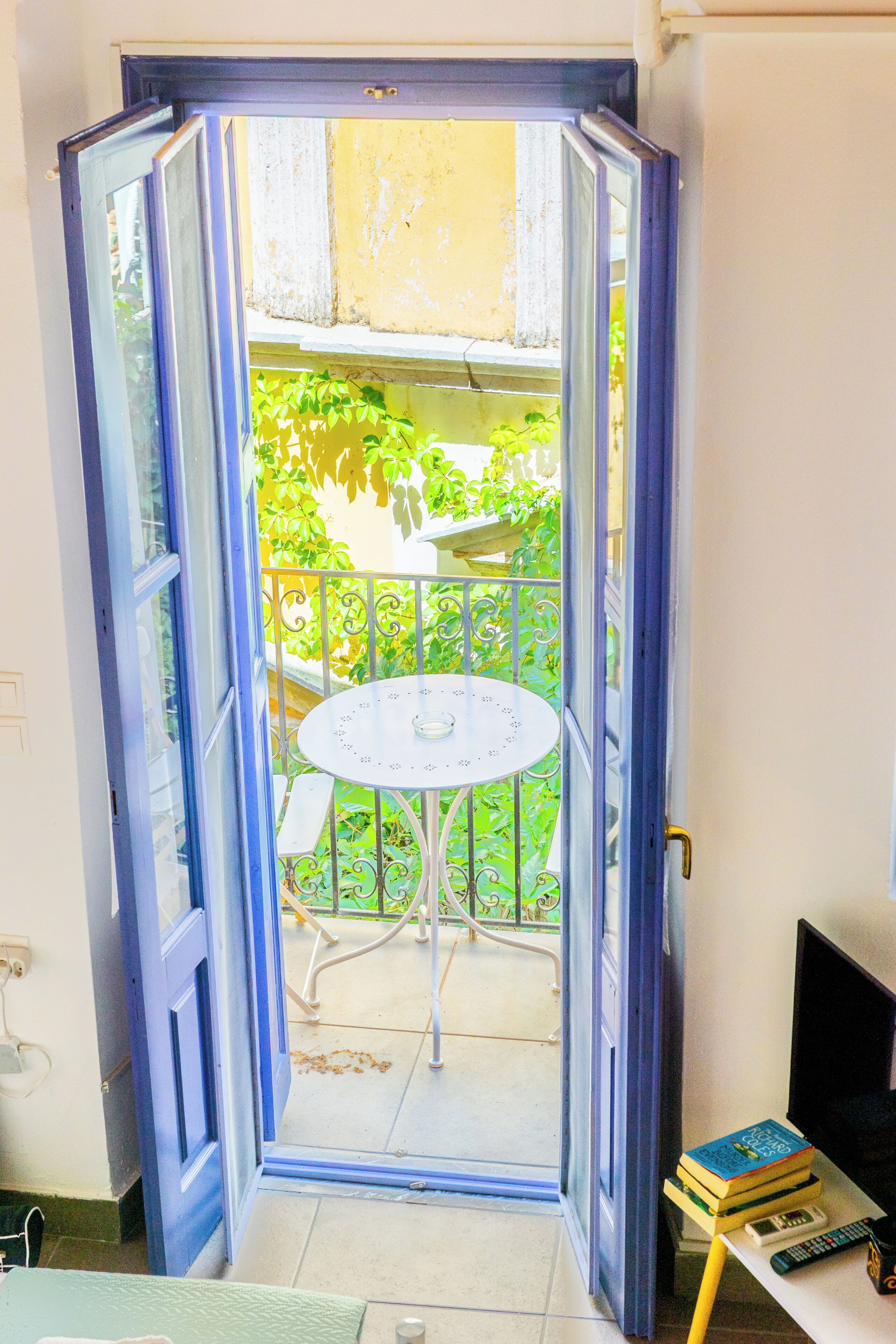 Puerta abierta que da a un balcón con mesa y sillas, un pequeño jardín y un escritorio con libros y un control remoto.