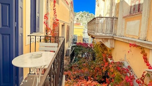 A narrow street with buildings on either side, a table with a glass on it, and a railing. - Kali Strata 2 - Downtown (Symi)