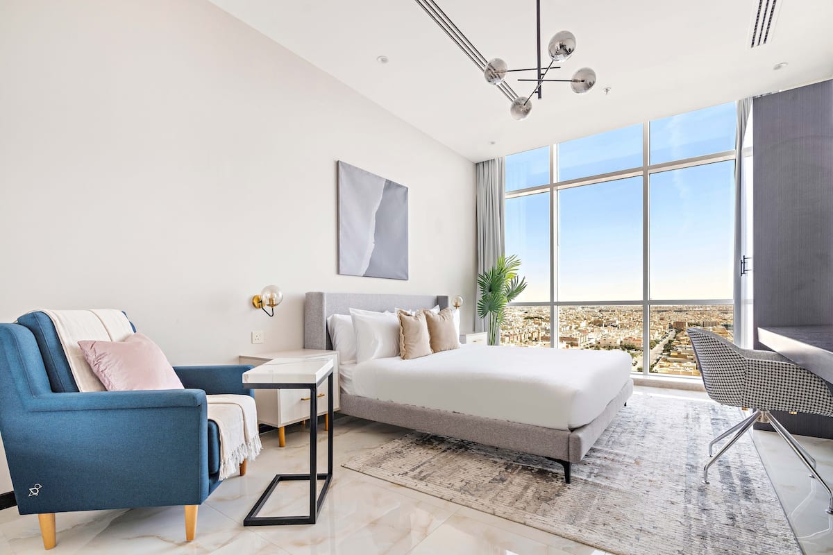 A modern bedroom with a large bed, a blue armchair, a small table, and a view of the city through large windows.