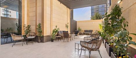 An outdoor seating area with wicker chairs and tables, surrounded by potted plants and a modern building facade.