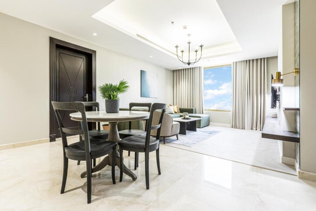 A modern dining area with a round table and chairs, a potted plant, and a view of the sky through a large window.