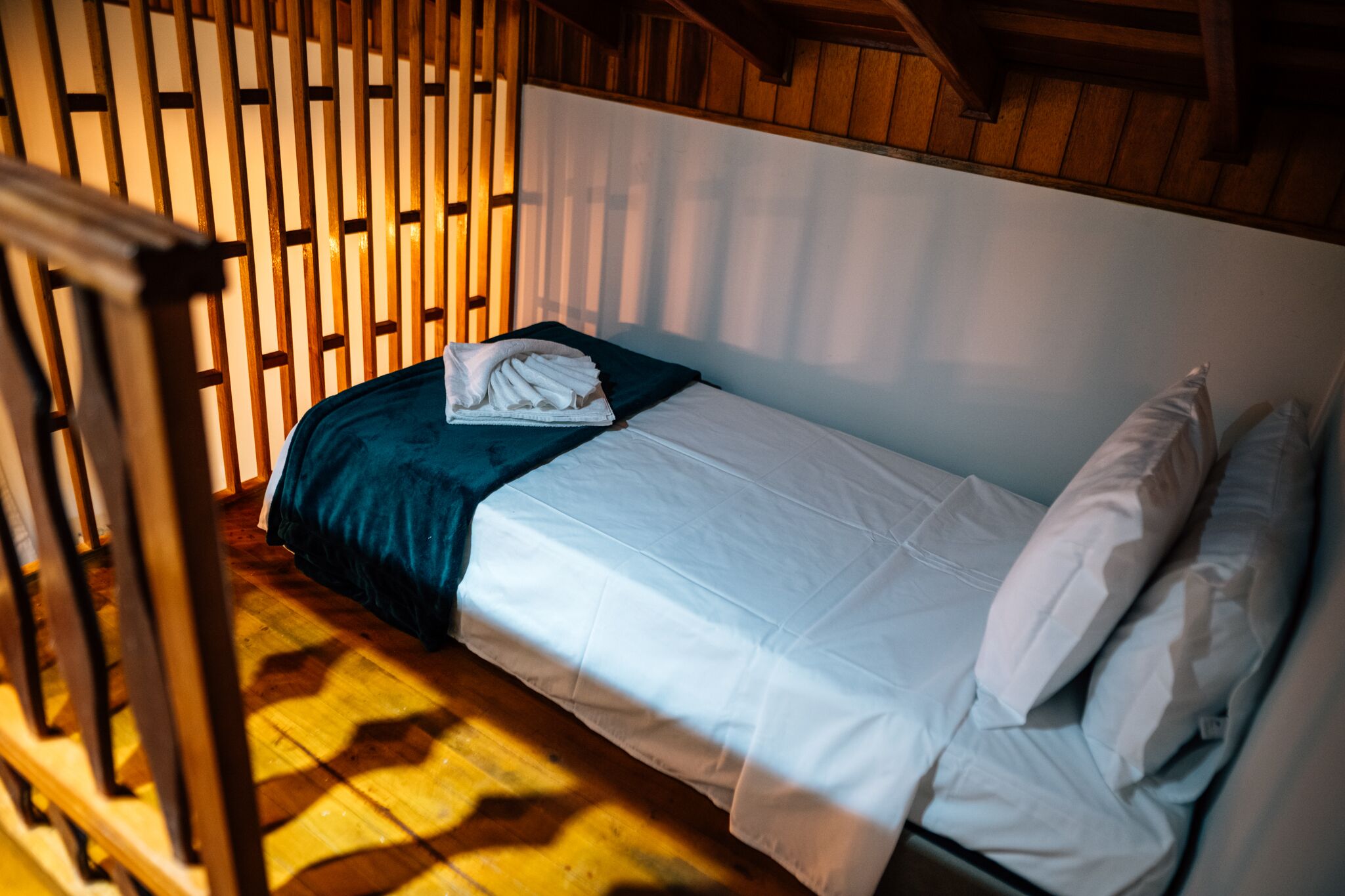 A single bed with white linens, a dark blue bedspread, and a pillow. The room has wooden walls and a slanted ceiling.
