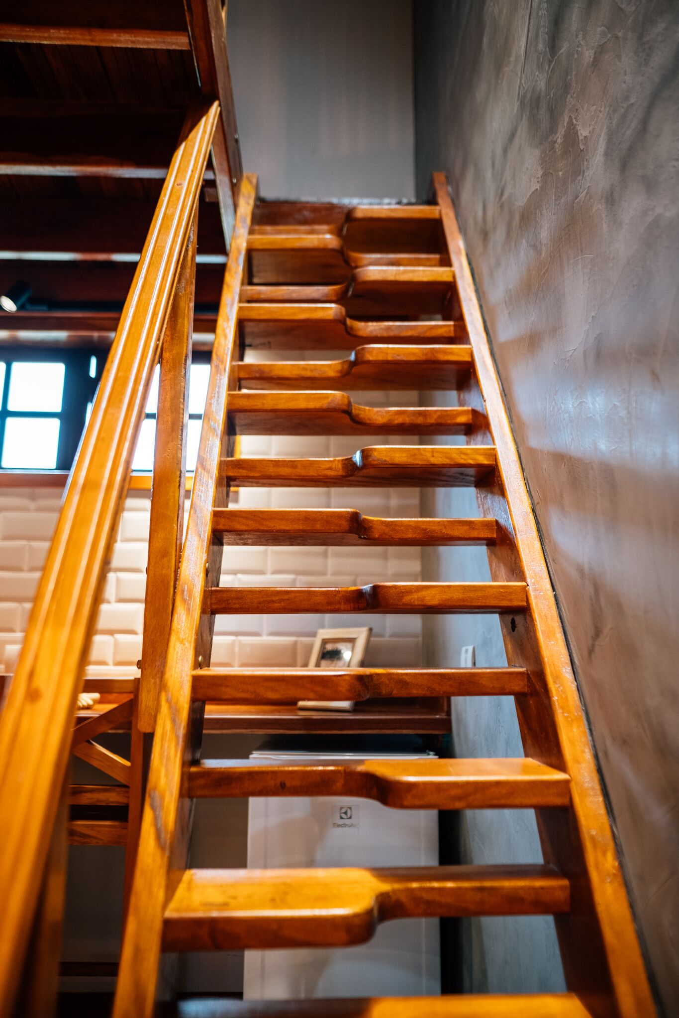 A wooden staircase with a handrail, leading up to a landing with a white wall and a window.