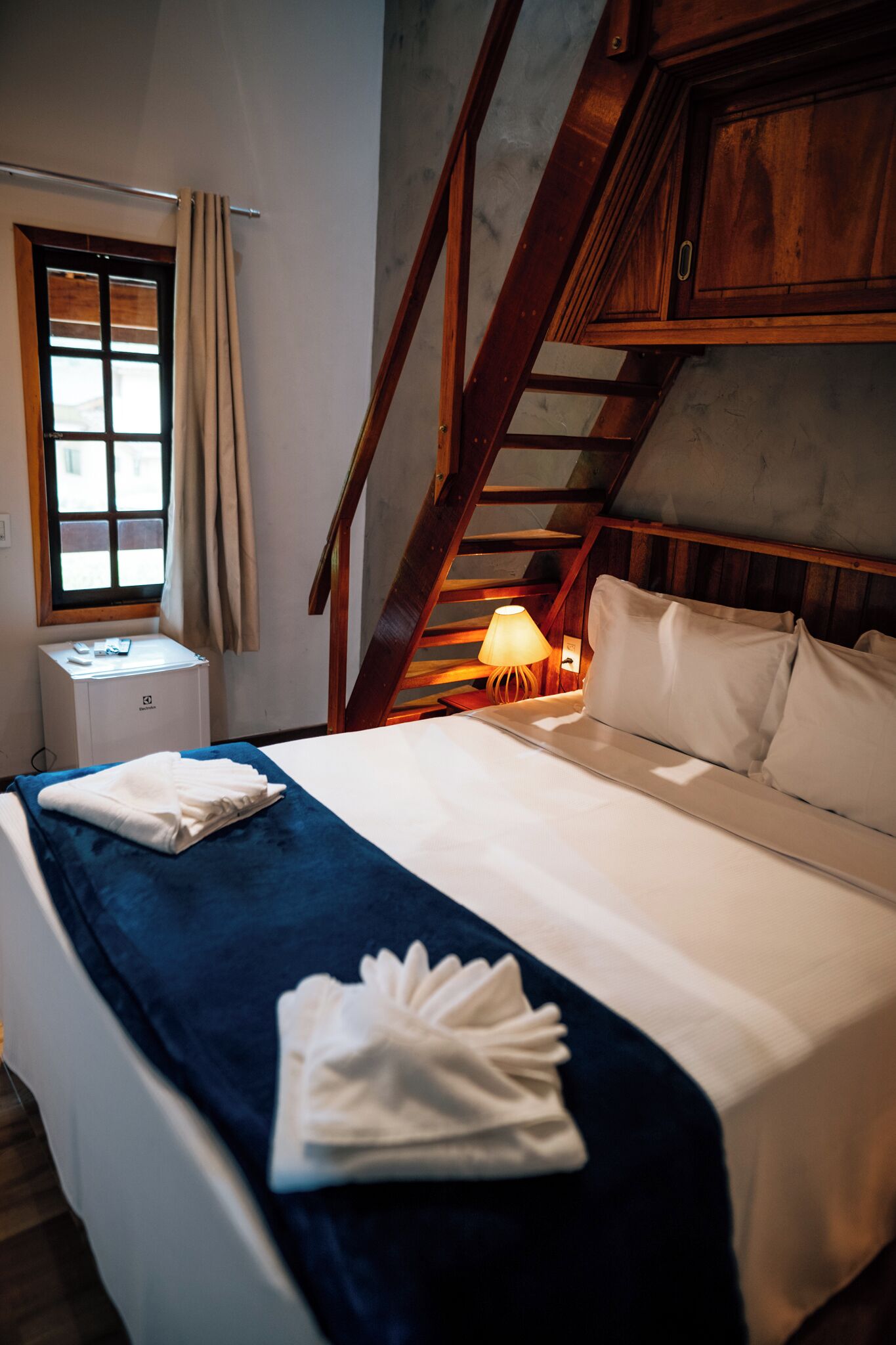 Habitación de hotel con cama, mesita de noche, lámpara, ventana con cortinas y una escalera de madera.