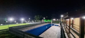 Vista nocturna de un área de piscina con plataforma de piedra, rodeada por una valla e iluminada con varias luces.