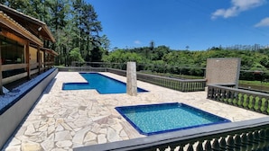 A pool area with a stone-tiled deck, a wooden deck with a railing, and a view of a forested hill. - Espaco Villa Verde  (Ibiuna)