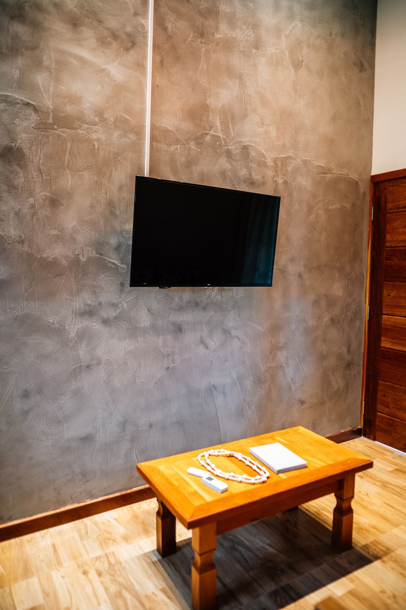 A room with a flat-screen TV mounted on a textured wall, a wooden table with a book and a white object, and a wooden cabinet.