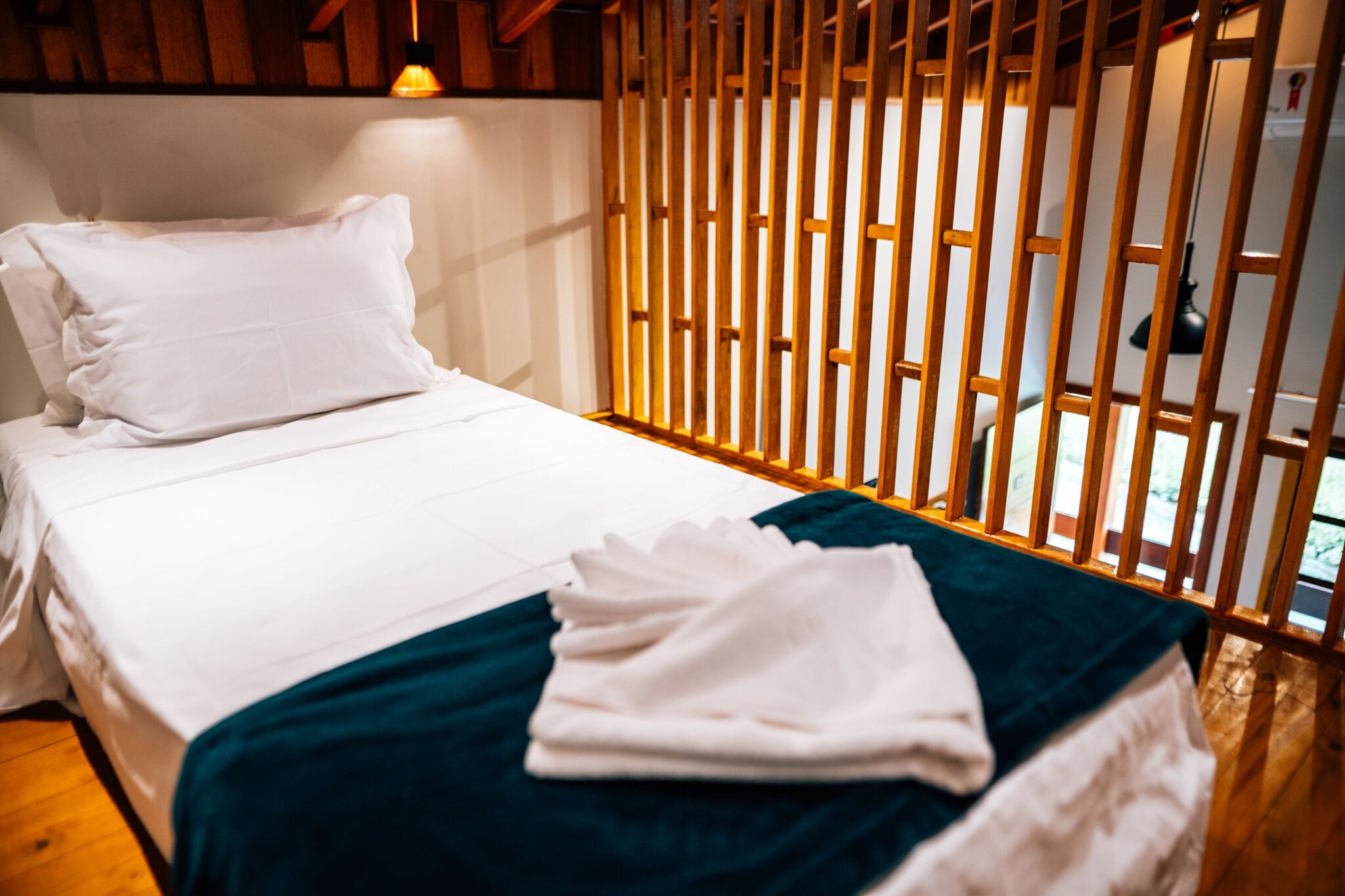 Un lit bien fait, avec du linge de lit blanc et un plaid turquoise, une cloison en lattes de bois et une lampe suspendue.