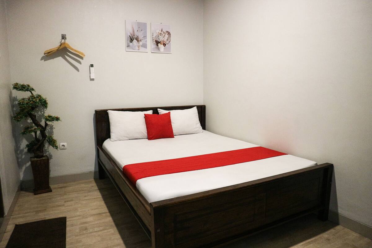 A neatly made bed with white and red bedding, a hanging garment rack, and a potted plant.