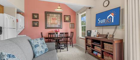 A living room with a gray sofa, wooden dining table, and chairs. There's a television on the wall and a ceiling fan.