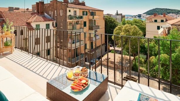 A balcony with a wicker table set with fruits and a book, overlooking a cityscape with buildings and trees. - Sun Spalato Harmony (Split)